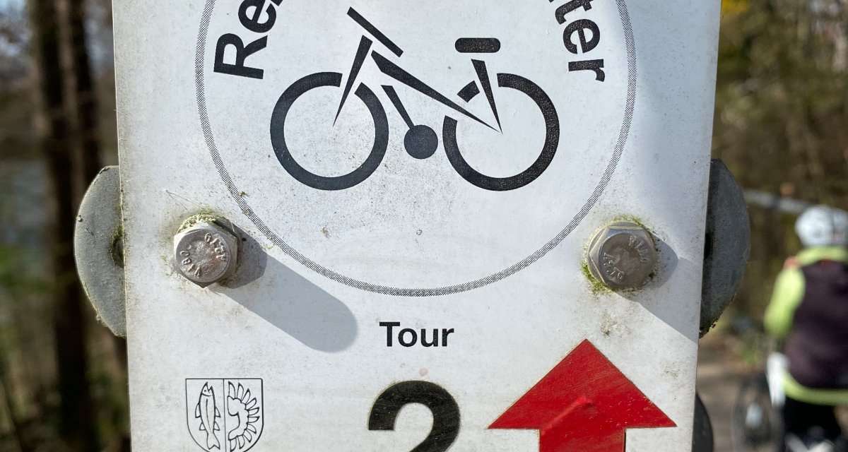 Schild mit 'Remseck-Achter Tour 2' und Fahrradsymbol an einem Pfosten, im Hintergrund ein Radfahrer auf einem Weg.