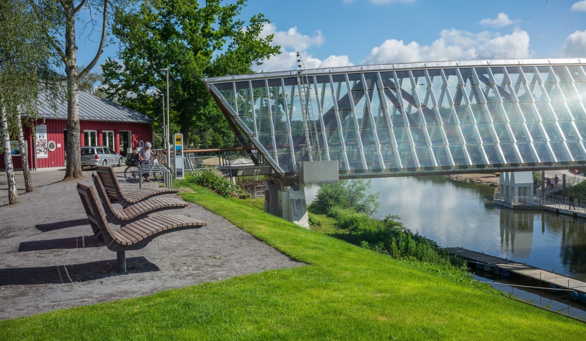 Moderne Glasbrücke über Fluss in Remseck am Neckar, umgeben von grüner Landschaft und einem roten Gebäude. Sitzbänke und Radfahrer im Vordergrund., © Remstal Tourismus e.V.