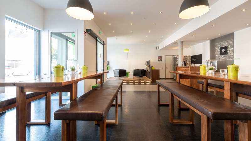 Moderne Lobby mit Holztischen und Bänken, großen Lampen und einem Sitzbereich im Hintergrund. Helle, einladende Atmosphäre., © a2 HOTELS Moderne Lobby mit Holztischen und Bänken, großen Lampen und einem Sitzbereich im Hintergrund. Helle, einladende Atmosphäre., © a2 HOTELS