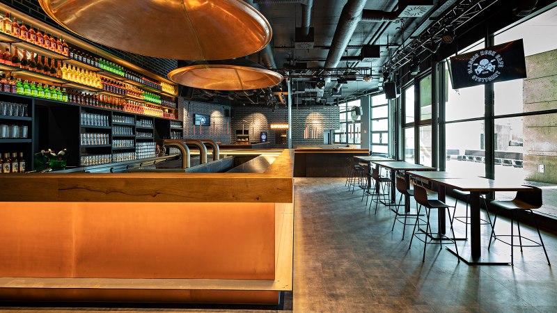 Modernes Caf&eacute; mit Bar, beleuchteten Regalen voller Flaschen, Gl&auml;sern und Sitzbereichen. Gro&szlig;e Fenster lassen viel Licht herein., &copy; Maks Richter