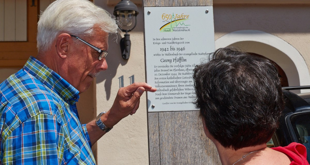 Zwei Personen lesen eine Gedenktafel an einem Holzpfosten. Die Tafel erinnert an historische Ereignisse in der Stadt Waldenbuch., © Natur.Nah. Schönbuch & Heckengäu