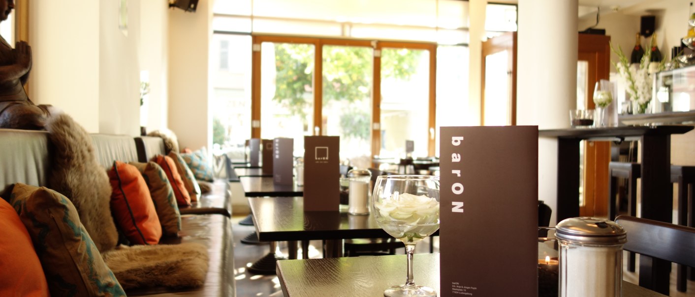 Gemütliches Café mit Kissen auf Bänken, Speisekarten auf Tischen und einem Glas mit Blüten. Große Fenster lassen viel Licht herein., © Andreas Ebert - BARON Gemütliches Café mit Kissen auf Bänken, Speisekarten auf Tischen und einem Glas mit Blüten. Große Fenster lassen viel Licht herein., © Andreas Ebert - BARON