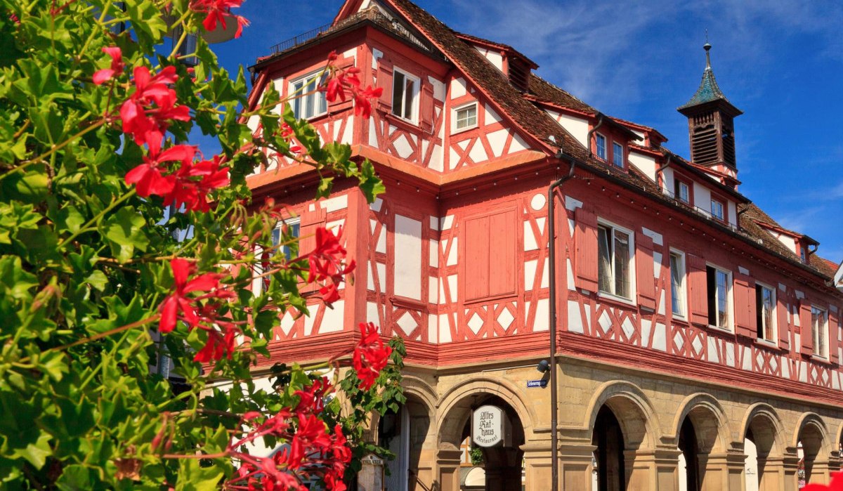 Das Alte Rathaus in Waiblingen, ein Fachwerkgebäude mit roten und weißen Details, umgeben von blühenden roten Blumen unter blauem Himmel., © WTM GmbH, Kai Koepf