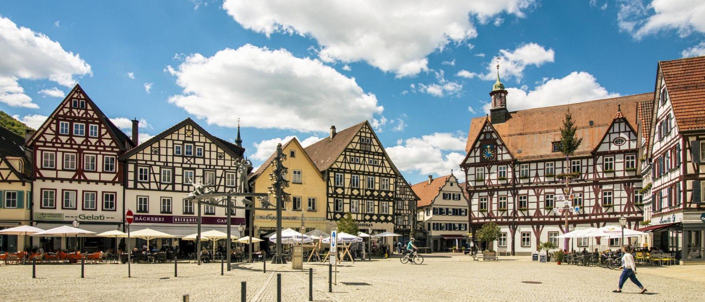 Marktplatz in Bad Urach mit malerischen Fachwerkhäusern, Cafés und Menschen bei sonnigem Wetter. Ein Radfahrer und Spaziergänger sind zu sehen., © Stuttgart-Marketing GmbH, Sarah Schmid Marktplatz in Bad Urach mit malerischen Fachwerkhäusern, Cafés und Menschen bei sonnigem Wetter. Ein Radfahrer und Spaziergänger sind zu sehen., © Stuttgart-Marketing GmbH, Sarah Schmid