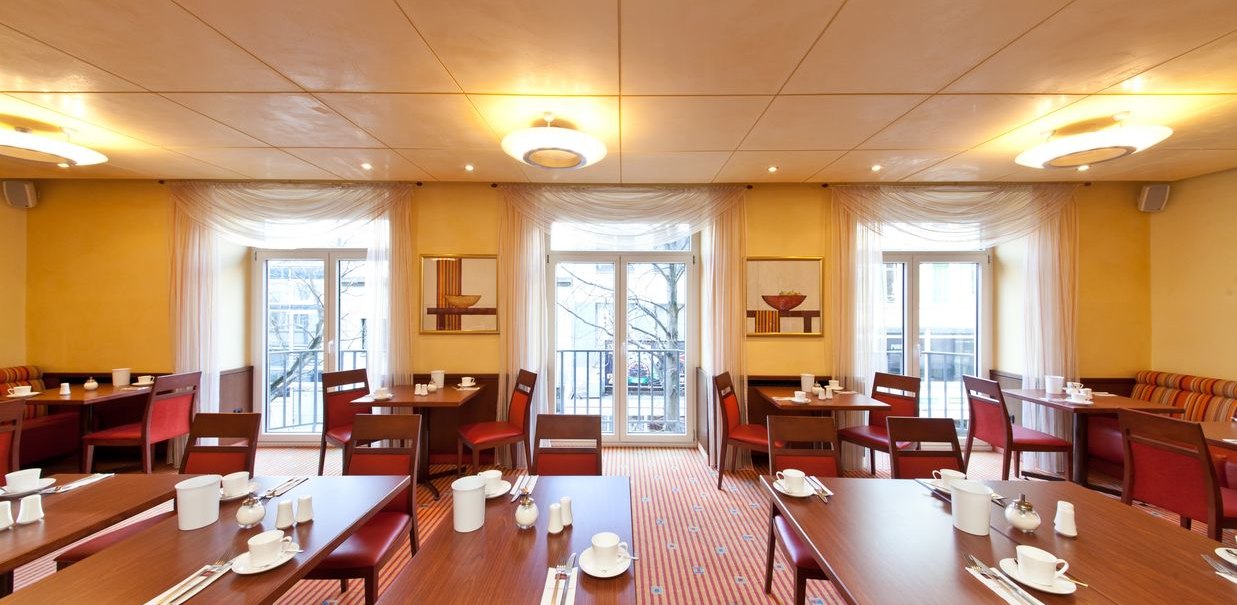 Ein heller Frühstücksraum mit gedeckten Tischen, roten Stühlen und großen Fenstern. Die Wände sind gelb, der Teppich rot gestreift., © Novum Hospitality