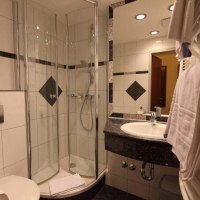 Modernes Badezimmer mit Glasdusche, Waschbecken und Handtuchhalter. Weiße Fliesen mit schwarzen Akzenten. Beleuchtung an der Decke., © Aparthotel Astro