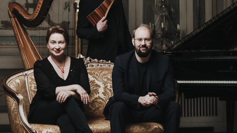 Drei Musiker sitzen und stehen in einem eleganten Raum mit einer Harfe und einem Klavier. Sie tragen formelle Kleidung und wirken konzentriert.