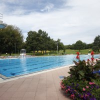 Ein Freibad mit einem großen Schwimmbecken, umgeben von grünen Bäumen und bunten Blumenbeeten. Der Himmel ist leicht bewölkt., © Stuttgarter Bäder