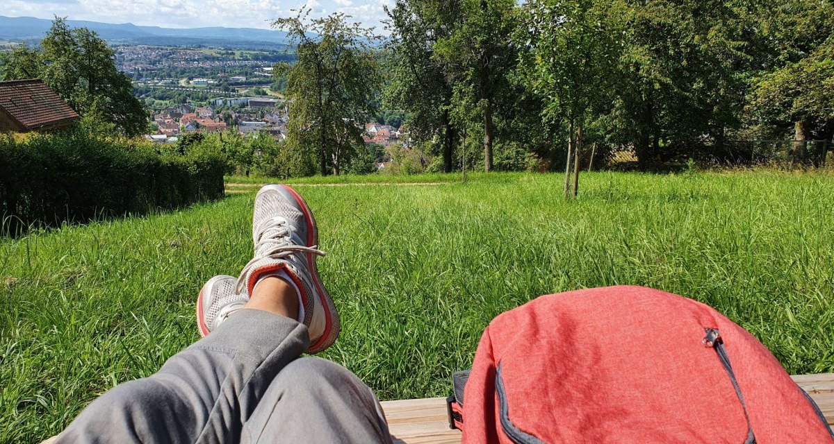 Person entspannt auf einer Bank mit Blick auf eine Stadt in der Ferne, umgeben von grünen Wiesen und Bäumen. Ein roter Rucksack liegt daneben. Person entspannt auf einer Bank mit Blick auf eine Stadt in der Ferne, umgeben von grünen Wiesen und Bäumen. Ein roter Rucksack liegt daneben.