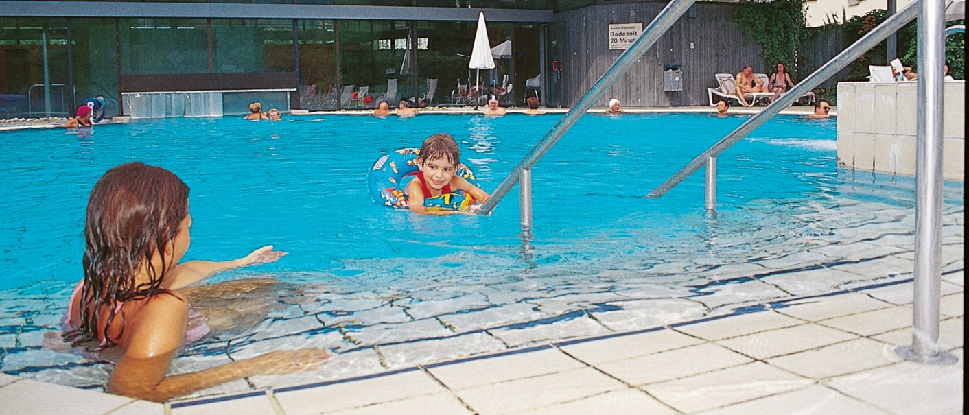 Ein Kind im Schwimmring im Pool, Erwachsene schwimmen und entspannen auf Liegestühlen im Badhaus Bad Boll., © Stuttgart-Marketing GmbH Ein Kind im Schwimmring im Pool, Erwachsene schwimmen und entspannen auf Liegestühlen im Badhaus Bad Boll., © Stuttgart-Marketing GmbH