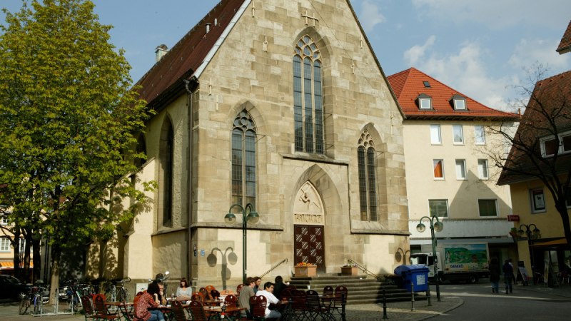 Die Nikolaikirche mit gotischen Fenstern, umgeben von Bäumen und Sitzgelegenheiten. Menschen sitzen draußen bei sonnigem Wetter., © Sphäre Verlag Die Nikolaikirche mit gotischen Fenstern, umgeben von Bäumen und Sitzgelegenheiten. Menschen sitzen draußen bei sonnigem Wetter., © Sphäre Verlag