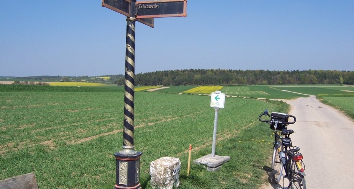 Ein historischer Wegweiser steht neben einem Feldweg. Ein Fahrrad lehnt an einem Schild. Im Hintergrund sind Felder und Wald zu sehen., © Natur.Nah. Schönbuch & Heckengäu Ein historischer Wegweiser steht neben einem Feldweg. Ein Fahrrad lehnt an einem Schild. Im Hintergrund sind Felder und Wald zu sehen., © Natur.Nah. Schönbuch & Heckengäu