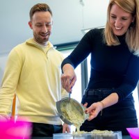 Zwei Personen gie&szlig;en gemeinsam Schokolade in eine Form. Beide l&auml;cheln und wirken konzentriert. Im Hintergrund sind Fenster zu sehen., &copy; Alfred Ritter GmbH &amp; Co. KG | Waldenbuch