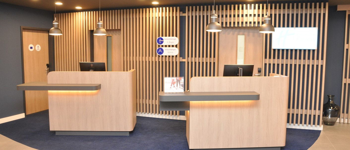 Moderne Rezeption im Holiday Inn Express Sindelfingen mit zwei Holztresen, blauer Teppich, hängende Lampen und Holzverkleidung an der Wand., © tristar GmbH Moderne Rezeption im Holiday Inn Express Sindelfingen mit zwei Holztresen, blauer Teppich, hängende Lampen und Holzverkleidung an der Wand., © tristar GmbH