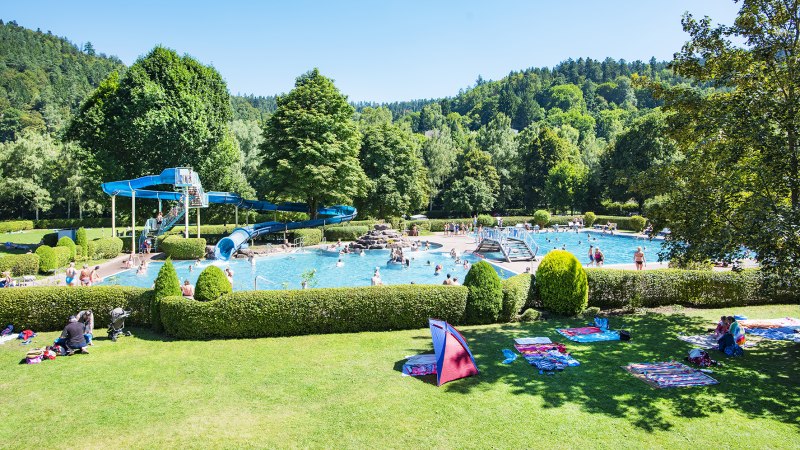 Freibad Bad Liebenzell mit blauer Rutsche, Schwimmern im Wasser und grüner Umgebung. Menschen entspannen auf der Wiese unter blauem Himmel., © LOCHER Fotodesing & Manufaktur Freibad Bad Liebenzell mit blauer Rutsche, Schwimmern im Wasser und grüner Umgebung. Menschen entspannen auf der Wiese unter blauem Himmel., © LOCHER Fotodesing & Manufaktur