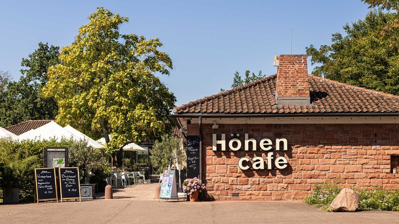 Das H&ouml;hencaf&eacute; im Killesbergpark zeigt ein Geb&auml;ude aus Backstein mit Terrasse, umgeben von B&auml;umen und Pflanzen. Wei&szlig;e Sonnenschirme sind sichtbar., &copy; Stuttgart-Marketing GmbH, Sarah Schmid