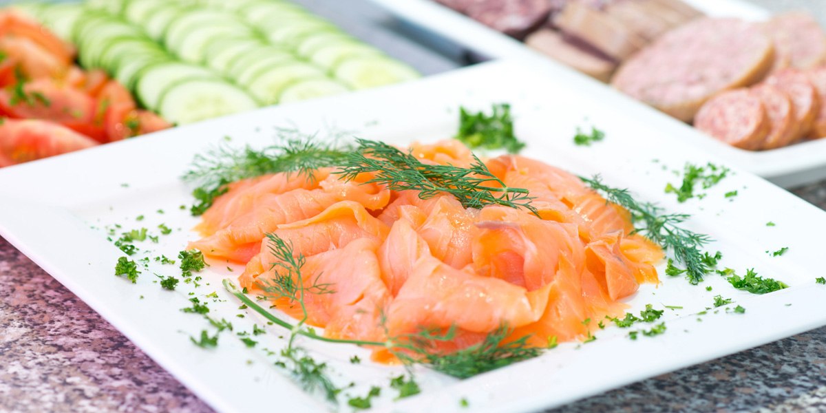 Geräucherter Lachs auf einem Teller, garniert mit Dill und Petersilie. Im Hintergrund Gurken- und Tomatenscheiben sowie Wurst., © ACHAT Hotels Deutschland