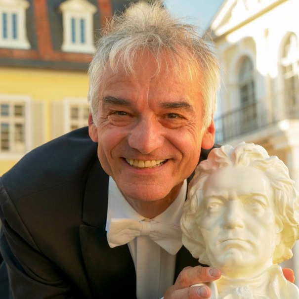 Ein lächelnder Mann in einem Anzug hält eine Beethoven-Büste vor einem historischen Gebäude mit gelber Fassade., © Stadtverwaltung Schorndorf