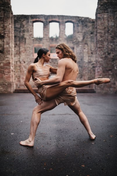 Zwei Balletttänzer in hautfarbenen Kostümen tanzen vor einer alten Steinruine. Eine Tänzerin wird elegant in die Luft gehoben., © Andrea D'Aquino