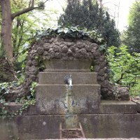 Ein alter Steinbrunnen mit Rosenverzierungen, umgeben von Efeu und B&auml;umen, steht auf einem Friedhof., &copy; cStadtN&uuml;rtingen