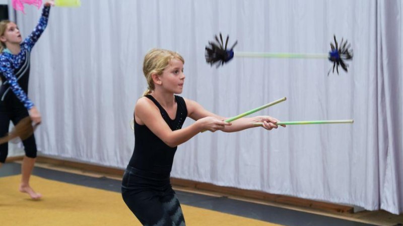 Kinder in einem Zirkus-Workshop jonglieren mit Stäben. Ein Mädchen im Vordergrund konzentriert sich auf ihre Übung. Hintergrund mit Vorhang., © Kunstschule Labyrinth