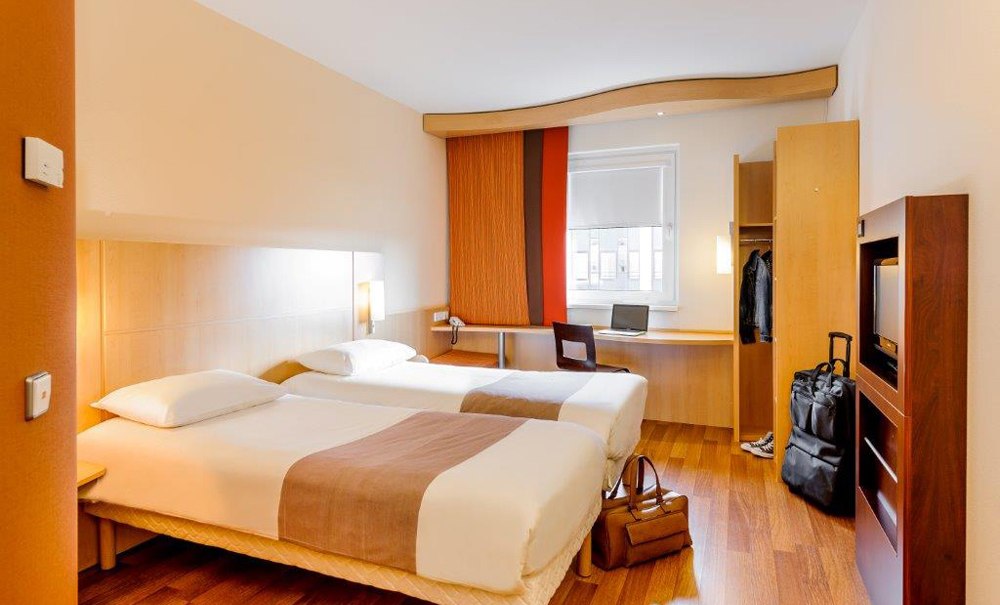 Ein Hotelzimmer mit zwei Einzelbetten, Schreibtisch mit Laptop, Kleiderschrank und Gepäck auf Holzboden., © Ibis Stuttgart Airport Messe Ein Hotelzimmer mit zwei Einzelbetten, Schreibtisch mit Laptop, Kleiderschrank und Gepäck auf Holzboden., © Ibis Stuttgart Airport Messe