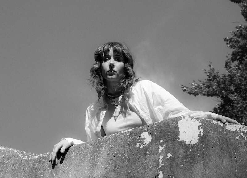 Schwarz-weiß Foto einer Person mit welligem Haar, die über eine Betonmauer schaut. Der Himmel ist klar, und Bäume sind im Hintergrund sichtbar., © Kulturhaus Schwanen Schwarz-weiß Foto einer Person mit welligem Haar, die über eine Betonmauer schaut. Der Himmel ist klar, und Bäume sind im Hintergrund sichtbar., © Kulturhaus Schwanen