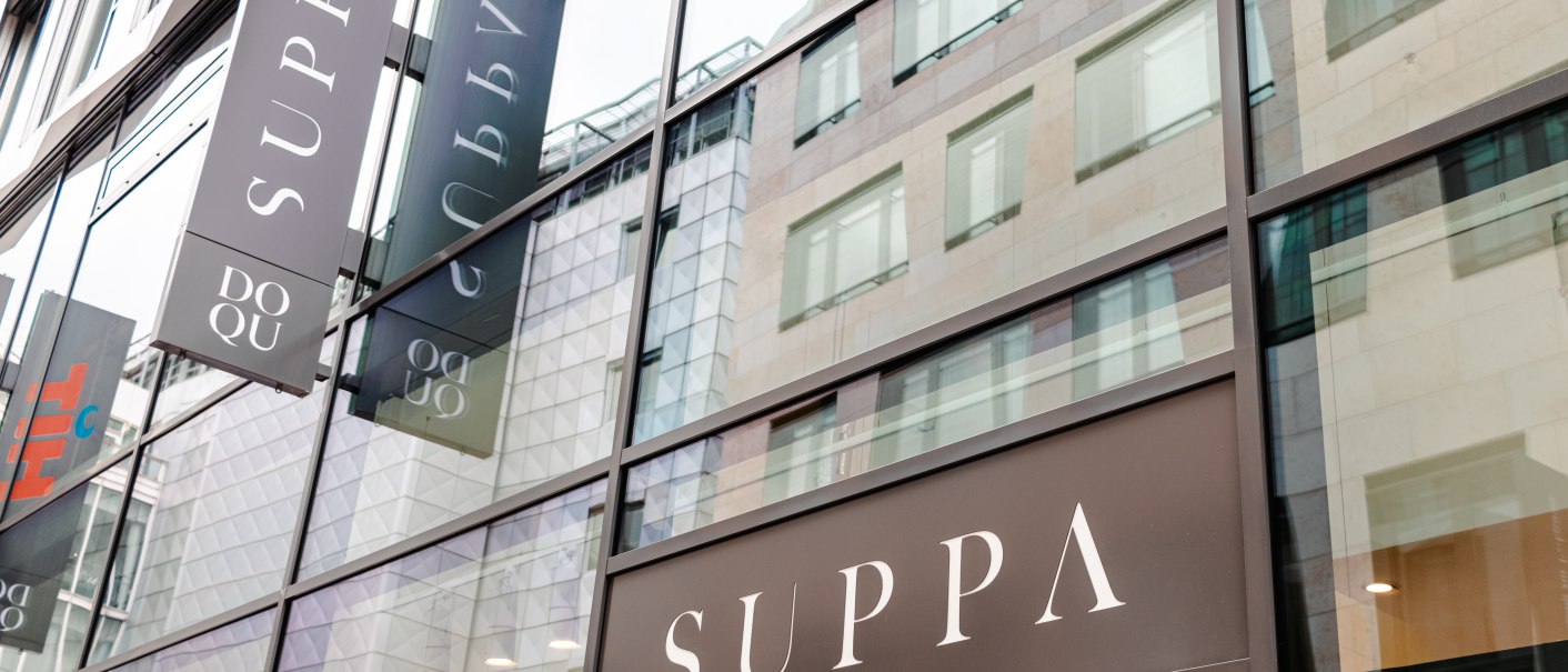 Glasfassade eines modernen Gebäudes mit mehreren SUPPA-Schildern. Die Spiegelung zeigt umliegende Architektur., © SMG_Sarah Schmid