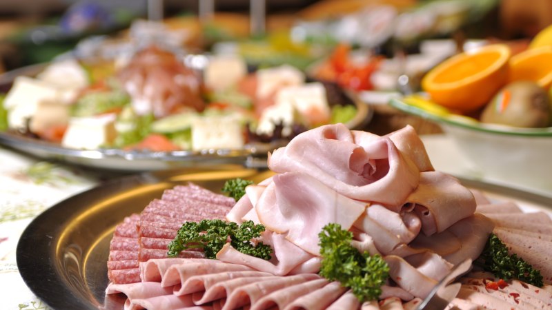 Ein Teller mit verschiedenen Wurstsorten, darunter Schinken und Salami, dekoriert mit Petersilie. Im Hintergrund sind weitere Speisen zu sehen., © TOMAS