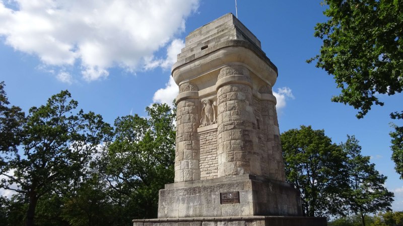 Ein massiver Bismarckturm aus Stein steht auf einem Platz, umgeben von Bäumen und unter einem blauen Himmel mit wenigen Wolken., © SMG Ein massiver Bismarckturm aus Stein steht auf einem Platz, umgeben von Bäumen und unter einem blauen Himmel mit wenigen Wolken., © SMG