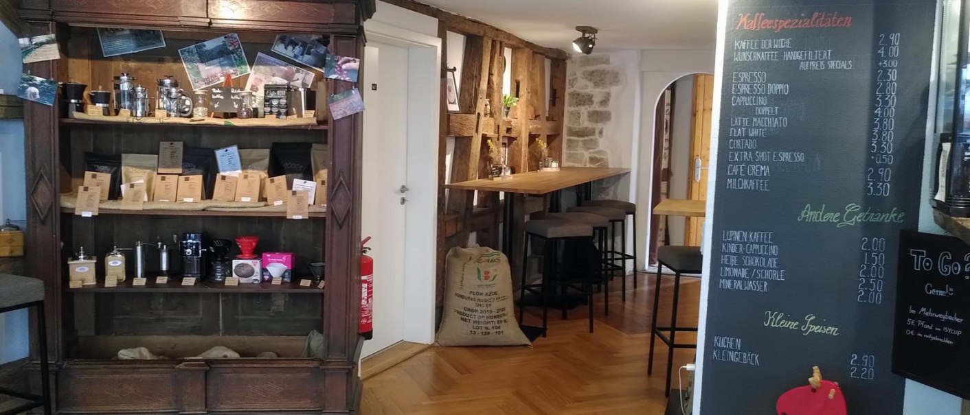 Kaffeerösterei mit Holzregal voller Kaffeeprodukte, Getränketafel und Sitzbereich. Gemütliche Atmosphäre mit Holzboden und Dekoration., © Matthias Gohl Kaffeerösterei mit Holzregal voller Kaffeeprodukte, Getränketafel und Sitzbereich. Gemütliche Atmosphäre mit Holzboden und Dekoration., © Matthias Gohl