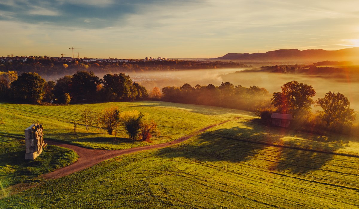 Sonnenaufgang über grünen Feldern mit Bäumen, einem Kunstwerk und Nebel in der Ferne. Die Sonne wirft lange Schatten., © Landratsamt Ostalbkreis Sonnenaufgang über grünen Feldern mit Bäumen, einem Kunstwerk und Nebel in der Ferne. Die Sonne wirft lange Schatten., © Landratsamt Ostalbkreis
