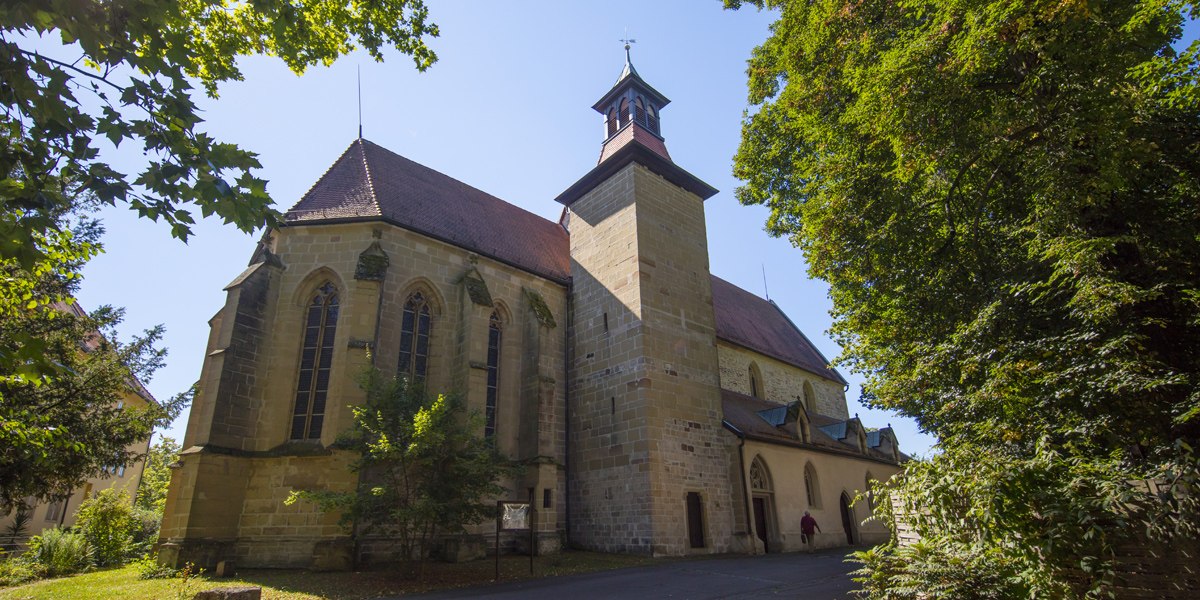 Die Schlosskirche Winnenden, ein historisches Gebäude mit gotischen Fenstern und einem Turm, umgeben von grünen Bäumen im Sonnenschein., © Stuttgart-Marketing GmbH Die Schlosskirche Winnenden, ein historisches Gebäude mit gotischen Fenstern und einem Turm, umgeben von grünen Bäumen im Sonnenschein., © Stuttgart-Marketing GmbH