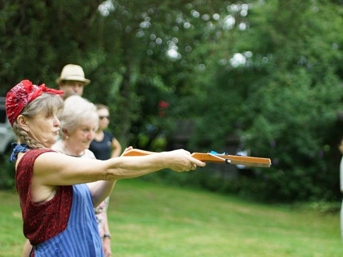Frau mit rotem Kopftuch zielt mit Holzgewehr im Freien, umgeben von anderen Personen. Grünfläche und Bäume im Hintergrund., © Cool-Tours StattReisen Frau mit rotem Kopftuch zielt mit Holzgewehr im Freien, umgeben von anderen Personen. Grünfläche und Bäume im Hintergrund., © Cool-Tours StattReisen