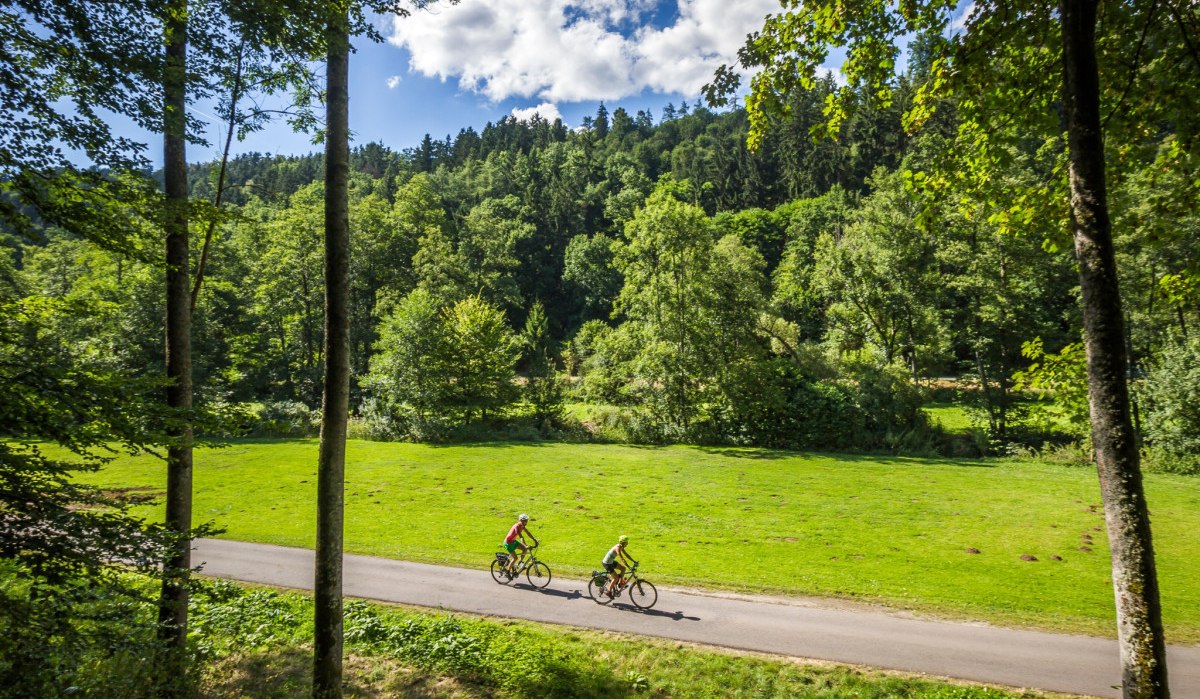 Zwei Radfahrer fahren auf einem asphaltierten Weg durch eine gr&uuml;ne Landschaft mit Wiesen und B&auml;umen, unter einem blauen Himmel mit wei&szlig;en Wolken., &copy; N&ouml;rdlicher Schwarzwald