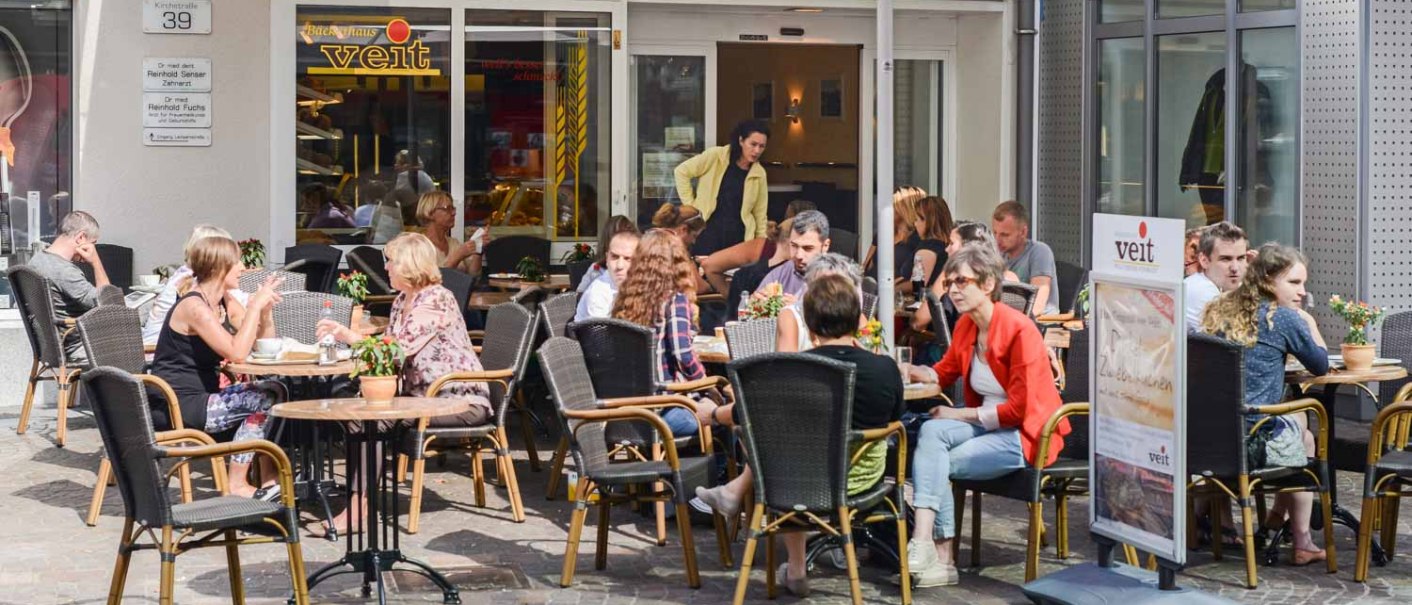 Menschen sitzen im Außenbereich der Bäckerei Veit in einer Fußgängerzone. Sie genießen das Wetter und unterhalten sich an Tischen unter Sonnenschirmen., © Bäckerei Veit, Dr. Michael Immendörfer