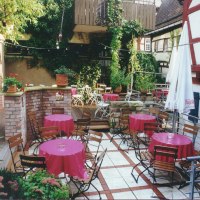 Ein gemütlicher Café-Innenhof mit Holztischen und -stühlen, rosa Tischdecken, umgeben von Pflanzen und Fachwerkhäusern., © 's Café