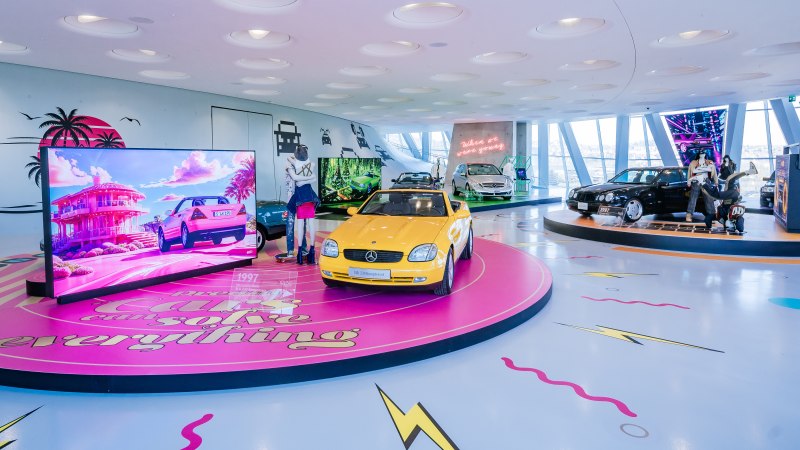 Bunte Ausstellung mit Youngtimer-Autos, darunter ein gelber Mercedes SLK. Neonlichter und Bildschirme mit Retro-Designs schm&uuml;cken den Raum., &copy; &copy; Mercedes-Benz AG | Foto: Thomas Niederm&uuml;ller