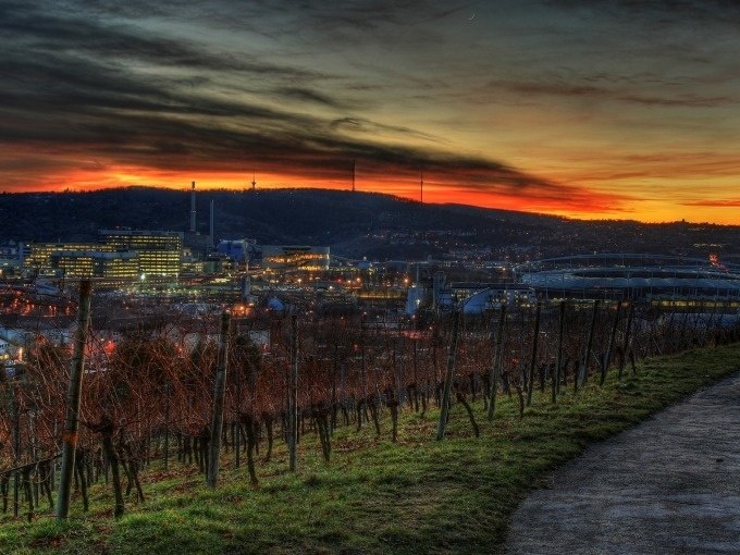 Abendlicher Blick auf eine Stadt mit beleuchteten Geb&auml;uden und einem Stadion, umgeben von Weinbergen. Der Himmel ist dramatisch rot-orange gef&auml;rbt., &copy; Cool-Tours StattReisen
