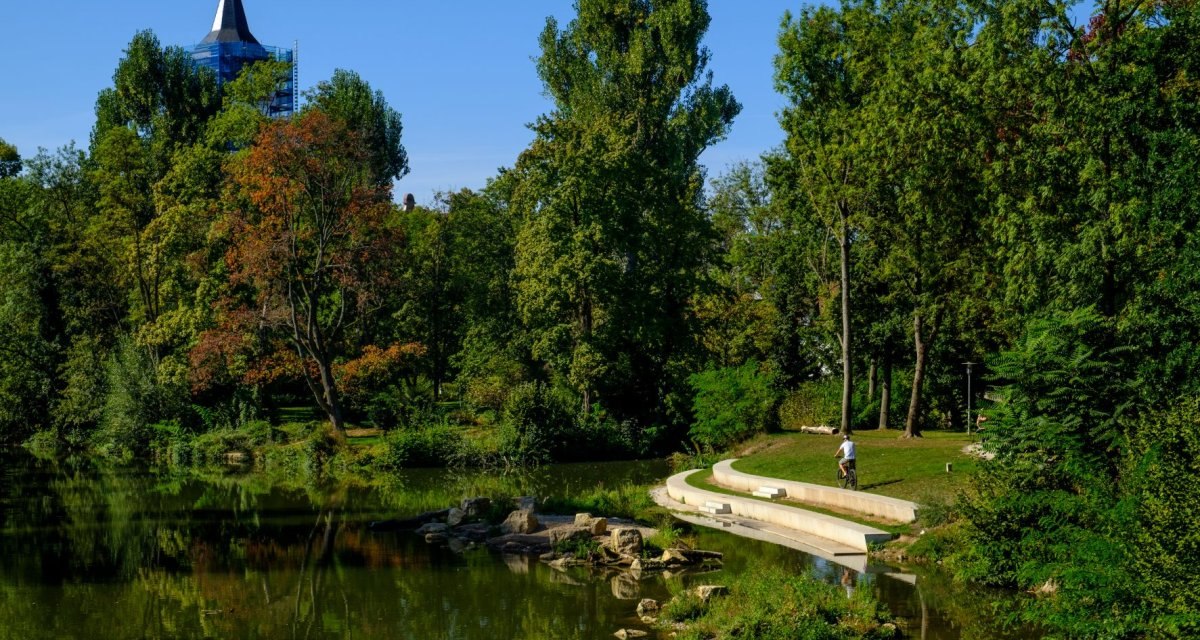 Ein grüner Park mit einem Teich, Bäumen und einem Turm im Hintergrund. Eine Person mit Fahrrad auf einem Weg., © WTM GmbH Waiblingen Ein grüner Park mit einem Teich, Bäumen und einem Turm im Hintergrund. Eine Person mit Fahrrad auf einem Weg., © WTM GmbH Waiblingen