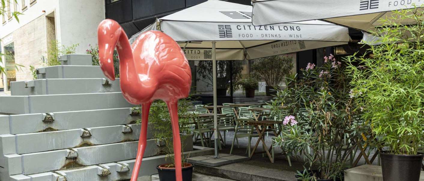 Ein großer, pinker Flamingo steht vor dem Café Citizen Long. Im Hintergrund sind Tische, Stühle und Pflanzen zu sehen., © SMG, Sarah Schmid Ein großer, pinker Flamingo steht vor dem Café Citizen Long. Im Hintergrund sind Tische, Stühle und Pflanzen zu sehen., © SMG, Sarah Schmid