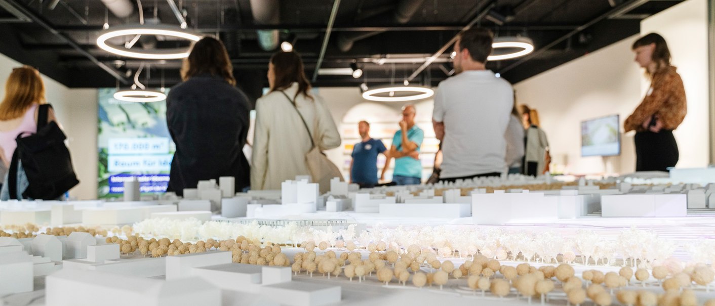Besucher betrachten ein Architekturmodell in einer Ausstellung. Moderne Deckenleuchten und ein Bildschirm im Hintergrund., © Dominique Brewing
