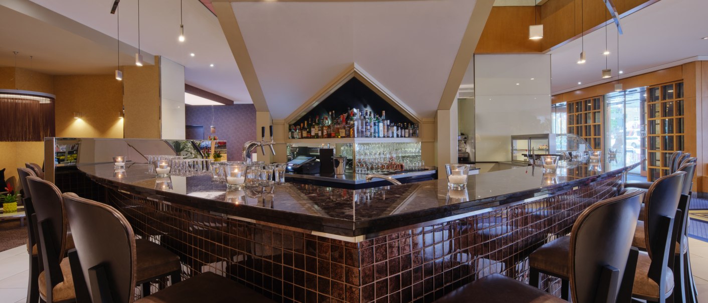 Moderne Bar mit dunklen Holzmöbeln, Hockern und stilvoller Beleuchtung. Flaschen und Gläser sind hinter der Theke sichtbar., © Steigenberger Graf Zeppelin