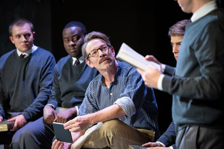 Schauspieler in Schuluniformen und ein Mann in Freizeitkleidung sitzen auf einer Bühne und lesen aus Büchern. Schauspieler in Schuluniformen und ein Mann in Freizeitkleidung sitzen auf einer Bühne und lesen aus Büchern.