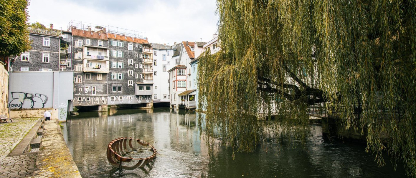 Ein malerischer Fluss flie&szlig;t durch Esslingen, ges&auml;umt von Fachwerkh&auml;usern und einer gro&szlig;en Trauerweide. Ein Kunstwerk ragt aus dem Wasser., &copy; SMG, Sarah Schmid