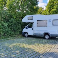 Ein Wohnmobil steht auf einem gepflasterten Stellplatz neben einer Ladestation, umgeben von Bäumen und einem Gebäude im Hintergrund., © Stadt Schorndorf