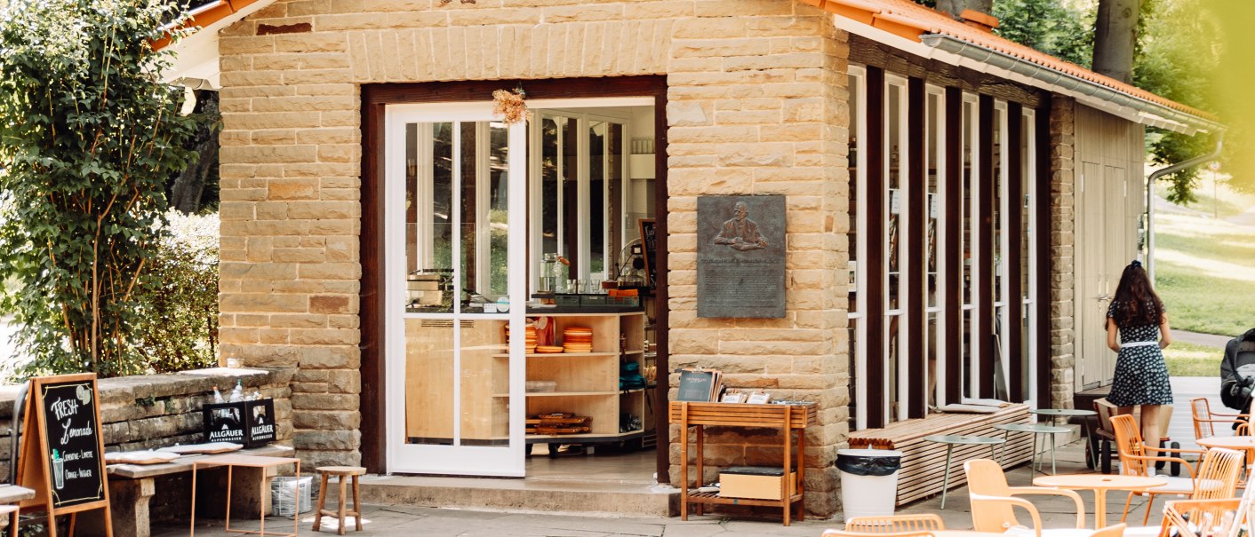 Kleines Café mit Steinwänden und großen Fenstern, umgeben von Bäumen. Orangefarbene Stühle und Tische auf der Terrasse, einladende Atmosphäre., © L. Prestle/ Documenting Love Kleines Café mit Steinwänden und großen Fenstern, umgeben von Bäumen. Orangefarbene Stühle und Tische auf der Terrasse, einladende Atmosphäre., © L. Prestle/ Documenting Love