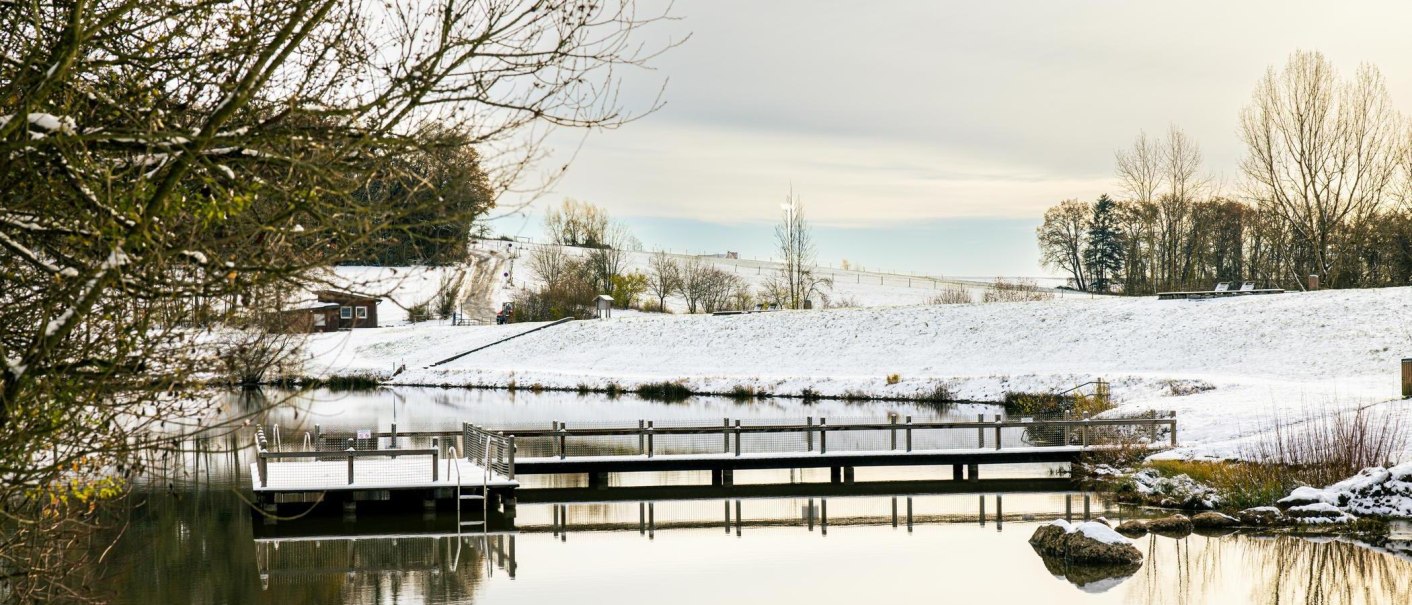 Winterliche Szene am Aichstruter Stausee mit schneebedecktem Steg und ruhigem Wasser. Im Hintergrund sind schneebedeckte Hügel und Bäume zu sehen., © Stuttgart-Marketing GmbH, Sarah Schmid Winterliche Szene am Aichstruter Stausee mit schneebedecktem Steg und ruhigem Wasser. Im Hintergrund sind schneebedeckte Hügel und Bäume zu sehen., © Stuttgart-Marketing GmbH, Sarah Schmid