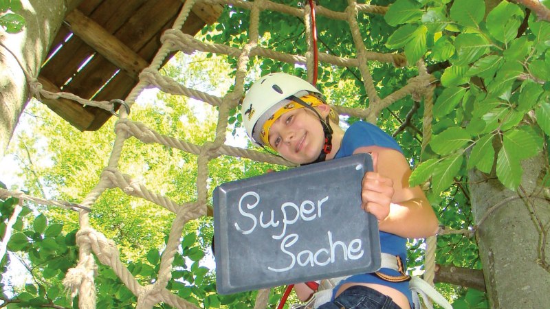 Person im Kletterpark hält eine Tafel mit der Aufschrift 'Super Sache' und trägt einen Helm. Im Hintergrund sind Bäume und ein Kletternetz zu sehen., © Stuttgart-Marketing GmbH