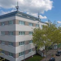Mehrst&ouml;ckiges Geb&auml;ude mit Antennen auf dem Dach, umgeben von B&auml;umen und parkenden Autos. Der Himmel ist leicht bew&ouml;lkt., &copy; Centro Hotels